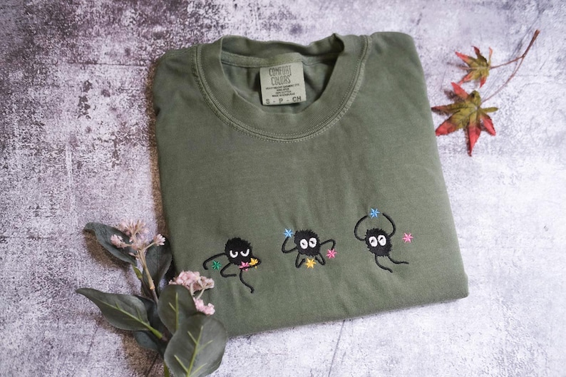 a green t-shirt with three small black figures and flowers embroidered on it, along with some leaves and flowers on a textured surface.