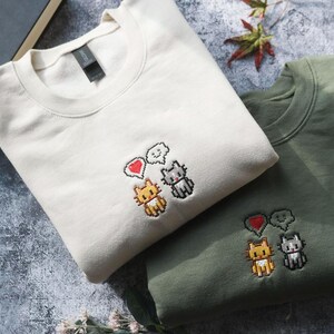 two t-shirts with cat designs, one white and one green, placed on a surface with some books and leaves.