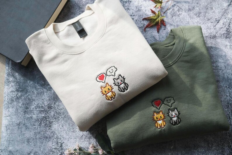 two t-shirts with cat designs, one white and one green, placed on a surface with some books and leaves.