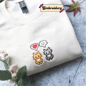 a white t-shirt with a pixel art design of three cartoon cats, each with a heart symbol, and a small plant or flower on the side.