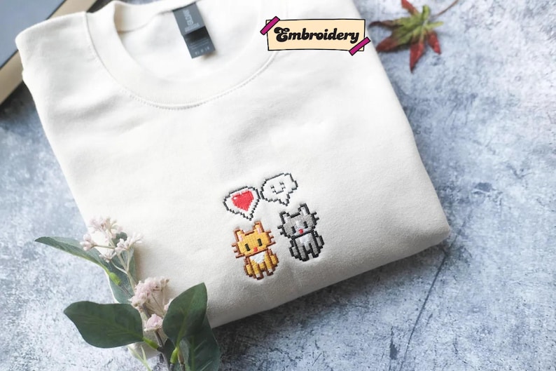 a white t-shirt with a pixel art design of three cartoon cats, each with a heart symbol, and a small plant or flower on the side.