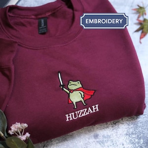 May include: Burgundy sweatshirt with an embroidered frog wearing a red cape and holding a sword. The word "Huzzah" is embroidered below the frog. The word "Embroidery" is in a white box.