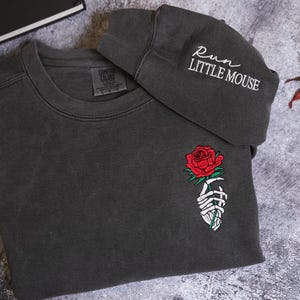 a gray sweatshirt with a rose embroidered on the front and a gray hat with the text "RUN LITTLE MOUSE" printed on it, placed on a gray surface with some leaves and flowers.