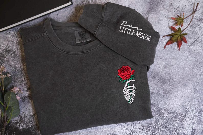 a gray sweatshirt with a rose embroidered on the front and a gray hat with the text "RUN LITTLE MOUSE" printed on it, placed on a gray surface with some leaves and flowers.