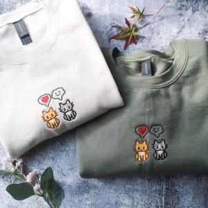 two t-shirts with cat embroidery, one white and one green, laid out on a gray surface.