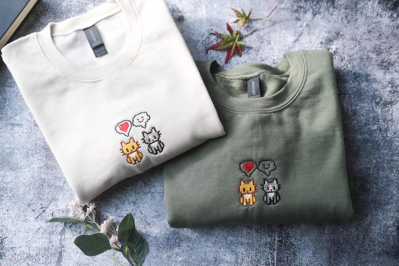 two t-shirts with cat embroidery, one white and one green, laid out on a gray surface.