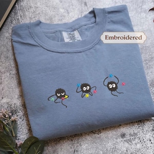 a blue t-shirt with three small black creatures with colorful eyes and arms, along with a label that says "Embroidered". The t-shirt is placed on a gray surface, and there are some leaves and a book visible in the background.