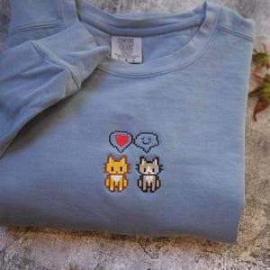 A gray sweatshirt with a pixelated design of two cats and a heart symbol on the front.