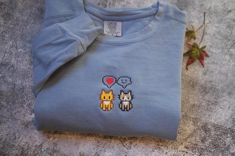 A gray sweatshirt with a pixelated design of two cats and a heart symbol on the front.