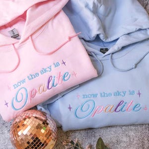 May include: Two hooded sweatshirts, one light pink and one light blue, with embroidered text that reads "now the sky is Opalite." The text is in a cursive font with a colorful gradient. A disco ball is in the foreground.