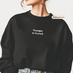 A person wearing a black sweater with the text " Enemies to lovers" embroidered on it.