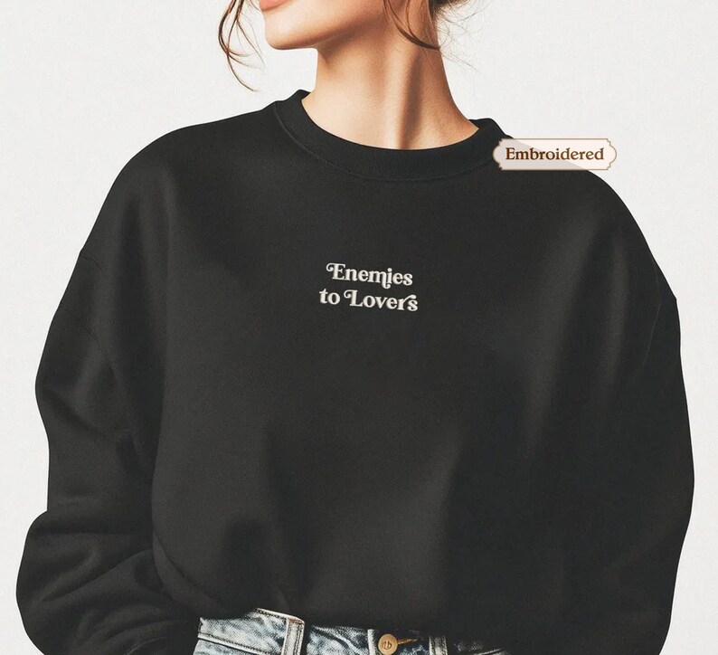 A person wearing a black sweater with the text " Enemies to lovers" embroidered on it.