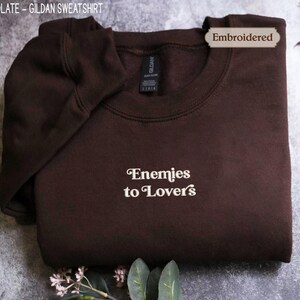 A dark brown hoodie with the text "Enemies to Lovers" printed on it, along with a label indicating that it is an embroidered item.
