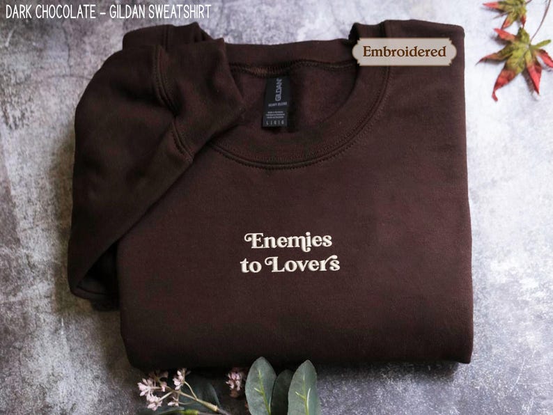 A dark brown hoodie with the text "Enemies to Lovers" printed on it, along with a label indicating that it is an embroidered item.