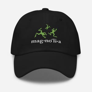 May include: Black baseball cap with embroidered green frog graphics and the word "mag·no'li·a" in white. The cap features a curved brim and an adjustable strap for a comfortable fit. A stylish accessory for casual wear.