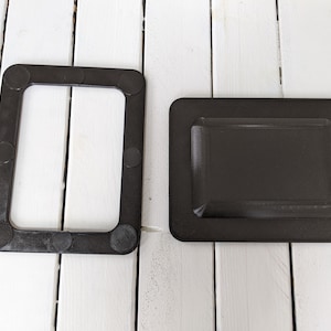 Op de afbeelding: Twee zwarte kunststof frames met afgeronde randen. Het frame aan de linkerkant heeft een rechthoekige opening en acht kleine zwarte cirkels rond de omtrek. Het frame aan de rechterkant is massief.