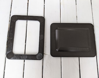 May include: Two black plastic frames with rounded edges. The frame on the left has a rectangular opening and eight small black circles around the perimeter. The frame on the right is solid.
