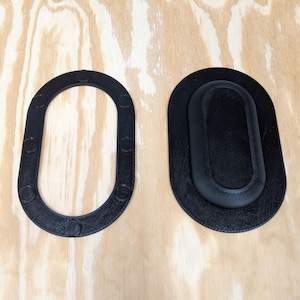 Puede incluir: Dos objetos negros de forma ovalada se muestran sobre una superficie de madera clara. Uno es un marco con hendiduras circulares, y el otro es un óvalo sólido y elevado dentro de un óvalo más grande. Los objetos parecen ser de plástico.