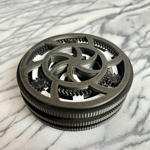 Pode incluir: Um brinquedo mecânico circular tipo fidget com um anel exterior cinzento-escuro e uma borda texturizada e estriada. A superfície superior apresenta um complexo mecanismo de engrenagens em camadas com um design em espiral central cinzento.