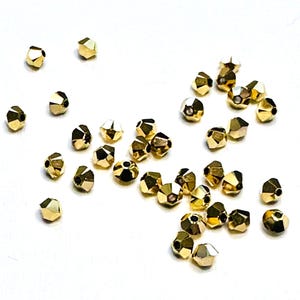 May include: Scattered on a white surface are numerous faceted gold beads. Each bead has multiple flat surfaces, reflecting light. The beads are small, likely used for jewelry making or crafting. The overall aesthetic is one of elegance and potential for creative projects.