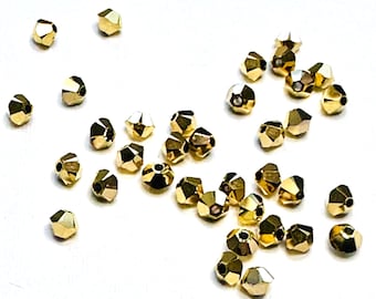 May include: Scattered on a white surface are numerous faceted gold beads. Each bead has multiple flat surfaces, reflecting light. The beads are small, likely used for jewelry making or crafting. The overall aesthetic is one of elegance and potential for creative projects.