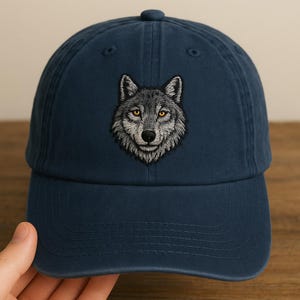 May include: A navy blue baseball cap featuring an embroidered wolf design. The wolf has detailed gray fur and yellow eyes. The cap has a curved brim and an adjustable strap for a comfortable fit. This hat is a stylish accessory.