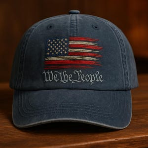 May include: A navy blue baseball cap featuring an embroidered American flag design with red and white stripes and stars. The phrase "We the People" is embroidered below the flag in a stylized font. The cap is made of a soft, worn material.