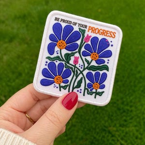 May include: A white patch with a floral design and the text "BE PROUD OF YOUR PROGRESS". The patch features blue flowers with orange centres and green stems. Pink buds are also present.