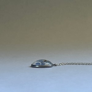 K&ouml;nnte beinhalten: Eine silberne Kettenkette mit einem runden Anh&auml;nger mit einem blauen und wei&szlig;en Design, das in klarem Harz gefasst ist.
