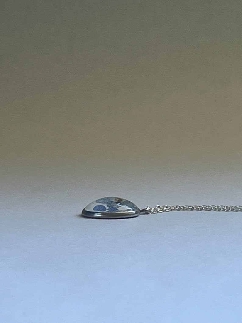 K&ouml;nnte beinhalten: Eine silberne Kettenkette mit einem runden Anh&auml;nger mit einem blauen und wei&szlig;en Design, das in klarem Harz gefasst ist.