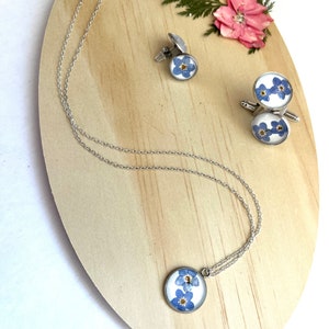 K&ouml;nnte beinhalten: Ein Set aus silbernem Schmuck mit Vergissmeinnicht-Blumen, die in klarem Harz gepresst sind. Das Set enth&auml;lt eine Halskette, ein Paar Manschettenkn&ouml;pfe und einen Anh&auml;nger.