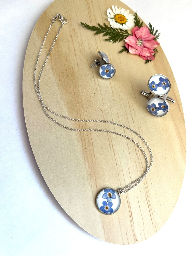 K&ouml;nnte beinhalten: Ein Set aus silbernem Schmuck mit Vergissmeinnicht-Blumen, die in klarem Harz gepresst sind. Das Set enth&auml;lt eine Halskette, ein Paar Manschettenkn&ouml;pfe und einen Anh&auml;nger.