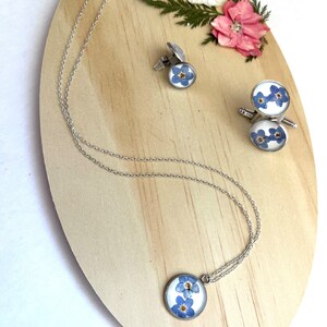 K&ouml;nnte beinhalten: Eine silberne Halskette, Manschettenkn&ouml;pfe und ein Paar Manschettenkn&ouml;pfe mit einem blauen Vergissmeinnicht-Blumendesign. Der Schmuck wird auf einer Holzoberfl&auml;che pr&auml;sentiert.