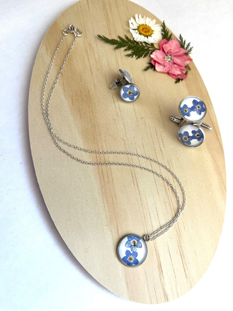 K&ouml;nnte beinhalten: Eine silberne Halskette, Manschettenkn&ouml;pfe und ein Paar Manschettenkn&ouml;pfe mit einem blauen Vergissmeinnicht-Blumendesign. Der Schmuck wird auf einer Holzoberfl&auml;che pr&auml;sentiert.