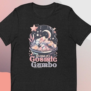 Könnte beinhalten: Dunkelgraues T-Shirt mit einer skurrilen Illustration einer kosmischen Gumbo-Szene. Das Design zeigt einen Halbmond, Sterne und eine Schüssel voller bunter, flauschiger Zutaten. Der Text auf dem Shirt lautet "It's kind of a Cosmic Gumbo."