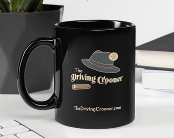 May include: Black ceramic mug with a curved handle. The mug features a graphic of a fedora hat and a cigar, with the text "The Driving Crooner" and "TheDrivingCrooner.com" in a vintage font. A small plant and a keyboard are in the background.