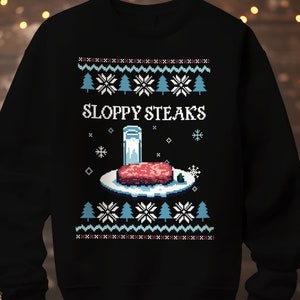 Può includere: Felpa nera con un design pixelato raffigurante un piatto con una bistecca, un bicchiere di latte e la scritta "SLOPPY STEAKS". Il design include fiocchi di neve, alberi di Natale e un motivo festivo, perfetto per le festività.