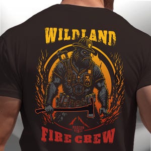 Può includere: Una maglietta marrone con una grafica di un pompiere con un casco e un'ascia. La grafica è circondata da fiamme e il testo "Wildland Fire Crew" è stampato sulla maglietta.
