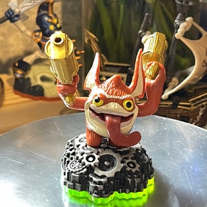 May include: A red and white figurine of a cartoon character with a long tongue, holding two gold-colored toy guns. The figure is mounted on a black base with silver gears and a neon green border. Other figurines are visible in the background.