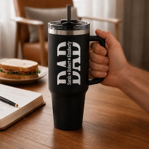 May include: A matte black stainless steel tumbler with a handle, a clear lid, and a straw. The front features the word DAD in large white letters, with the names Sarah and Kimberly separated by a star printed vertically between the letters.