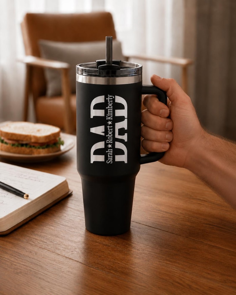 May include: A matte black stainless steel tumbler with a handle, a clear lid, and a straw. The front features the word DAD in large white letters, with the names Sarah and Kimberly separated by a star printed vertically between the letters.