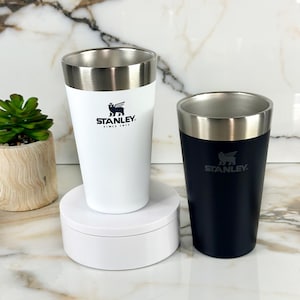May include: Two stainless steel Stanley insulated mugs, one white and one black, with the Stanley logo and "Since 1913" printed on them.
