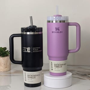 1 Black 2.0 30oz Stanley H2.0 Quencher Tumbler engraved with Ede Certified Public Accountants' logo next to 1 Lilac 30oz Stanley H2.0 Quencher Tumbler with the Stanley logo facing forward. The tumblers are set on a beautiful marble slab countertop.