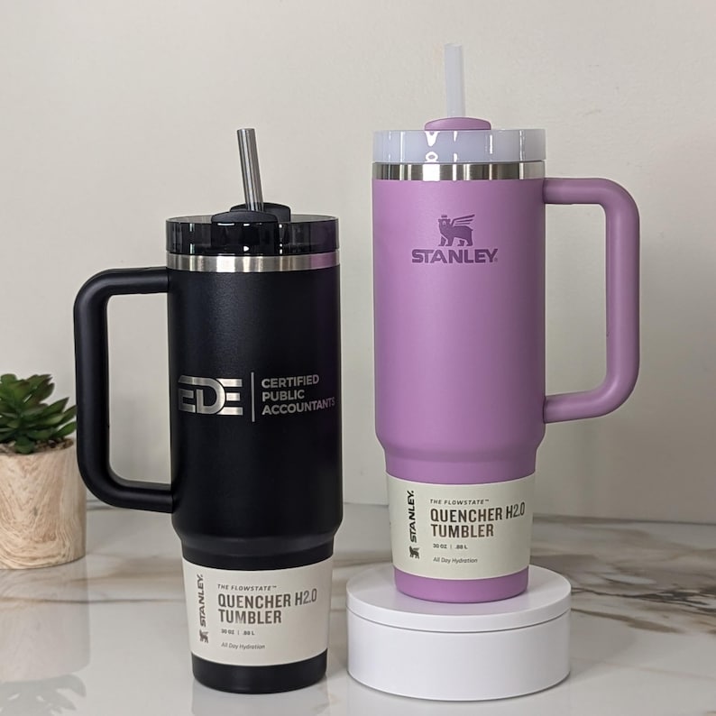 1 Black 2.0 30oz Stanley H2.0 Quencher Tumbler engraved with Ede Certified Public Accountants' logo next to 1 Lilac 30oz Stanley H2.0 Quencher Tumbler with the Stanley logo facing forward. The tumblers are set on a beautiful marble slab countertop.
