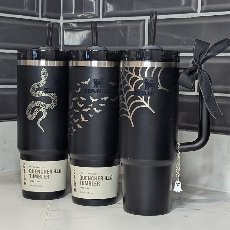 May include: Three matte black Stanley Quencher H2.0 tumblers with 30 oz capacity. The first features a silver snake, the second a swarm of bats, and the third a spiderweb design. The third tumbler has a black ribbon and a small ghost charm attached.
