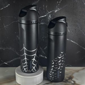 May include: Two matte black Owala water bottles with integrated carry loops and flip-top lids. The left bottle features a silver spider web graphic, while the right bottle displays a pattern of small silver bats. Both bottles have the Owala logo.
