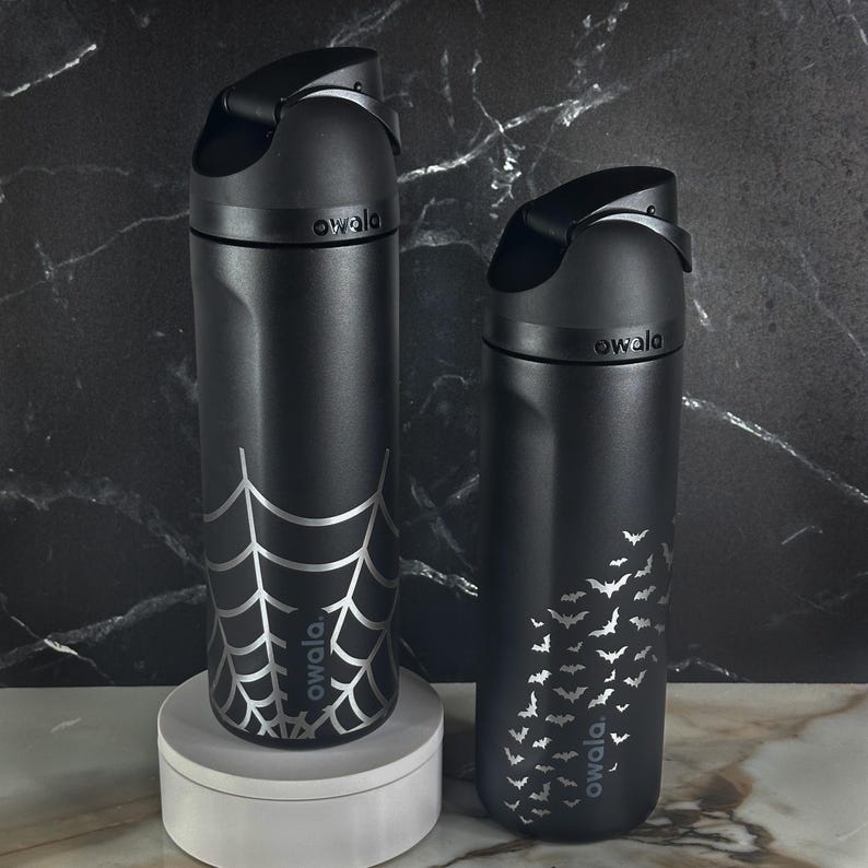 May include: Two matte black Owala water bottles with integrated carry loops and flip-top lids. The left bottle features a silver spider web graphic, while the right bottle displays a pattern of small silver bats. Both bottles have the Owala logo.