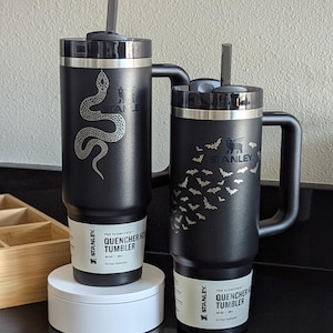 Two beautiful black 30oz Stanley Quencher tumblers in a kitchen setting. One tumbler features a shiny snake engraving that dynamically passes around the Stanley Logo. The other features an arrangement of bats that dynamically avoid the Stanley Logo.