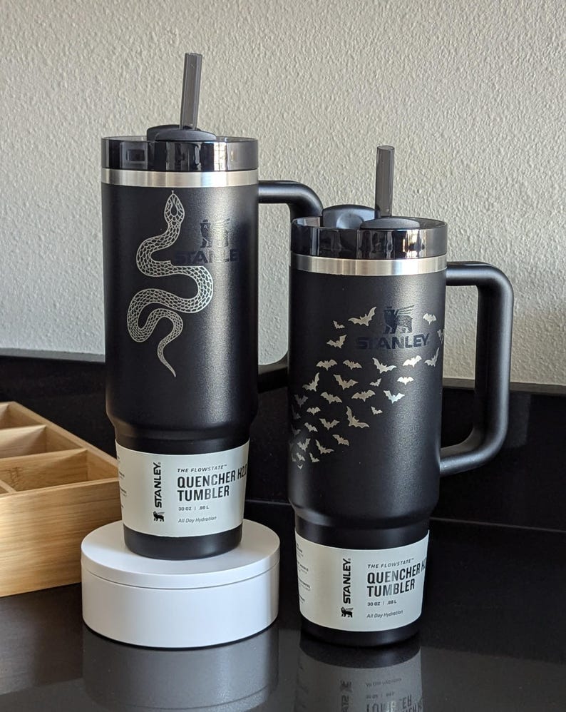 Two beautiful black 30oz Stanley Quencher tumblers in a kitchen setting. One tumbler features a shiny snake engraving that dynamically passes around the Stanley Logo. The other features an arrangement of bats that dynamically avoid the Stanley Logo.