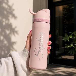 May include: A light pink Owala water bottle featuring a matte finish and a silver metallic script font reading Sophia vertically along the side. The bottle includes a matching pink lid with a built-in carry loop and a push-button release mechanism.